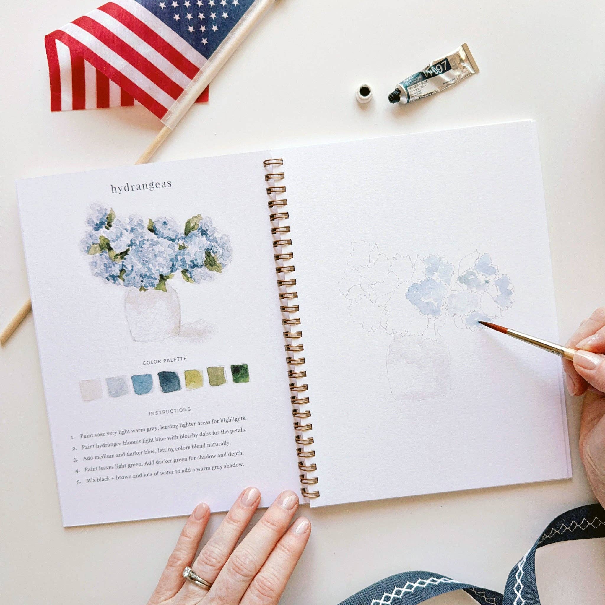 Open notebook with watercolor painting and color palette on a white surface with a small American flag.