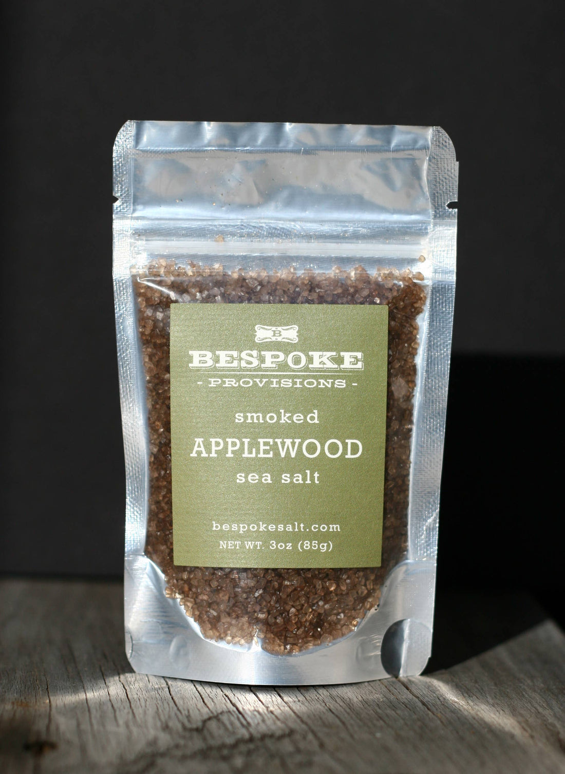A package of Bespoke Provisions Smoked Applewood Sea Salt placed on a wooden surface against a dark background.