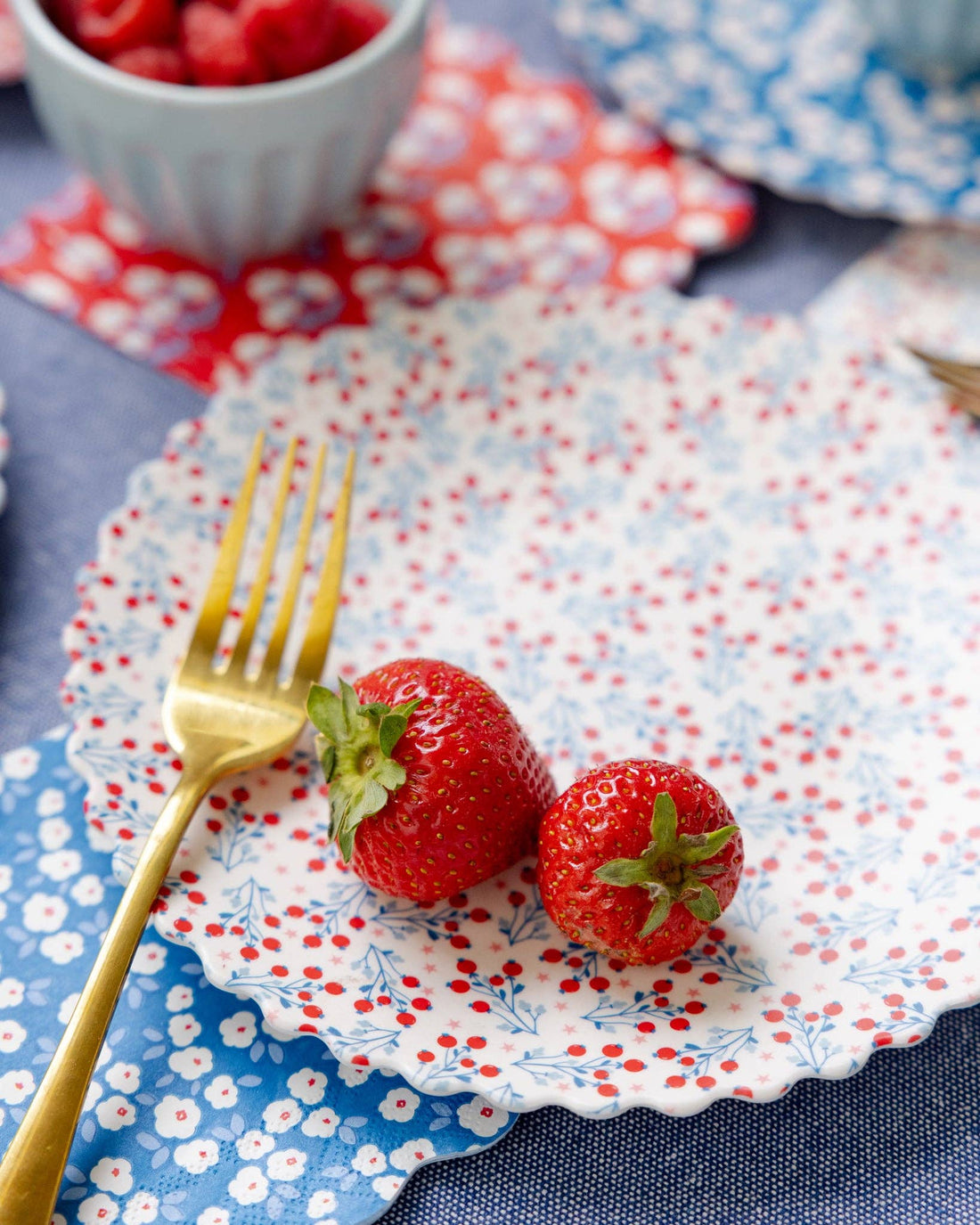 Two strawberries on a decorative plate with a gold fork, set against a floral tablecloth.