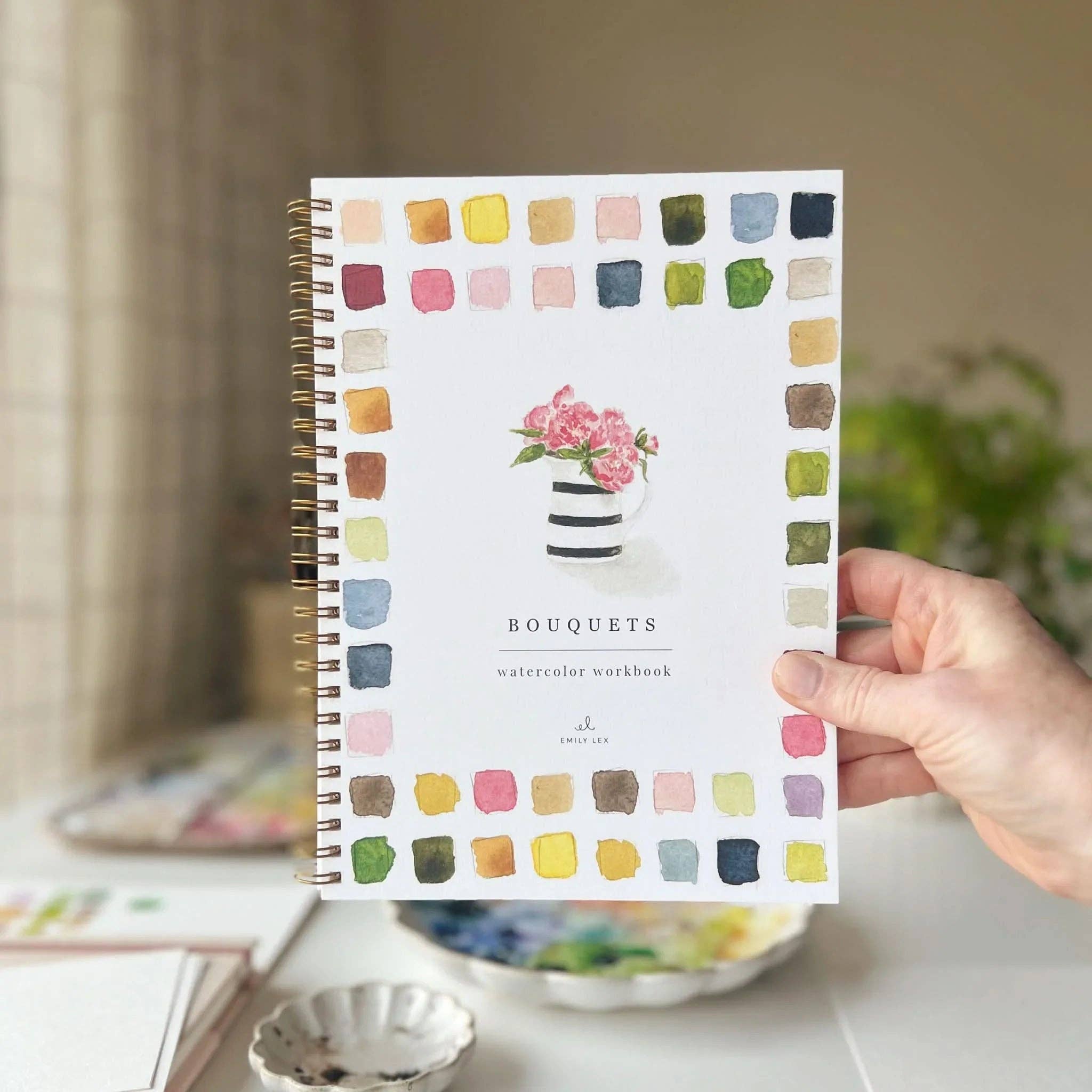 Hand holding a watercolor workbook with colorful square patterns on a blurred indoor background