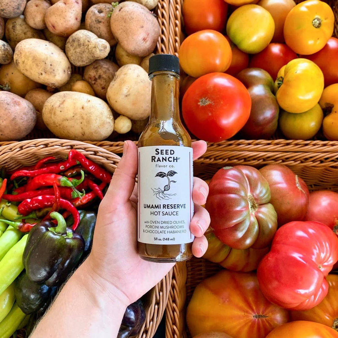 Hand holding a bottle of Seed Ranch Umami Reserve Hot Sauce amidst fresh vegetables.