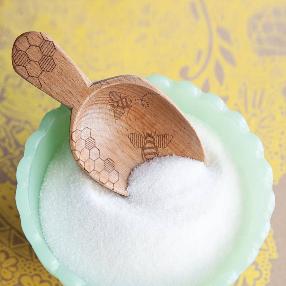 Wooden scoop with bee and honeycomb designs in a bowl of white powder on a yellow background