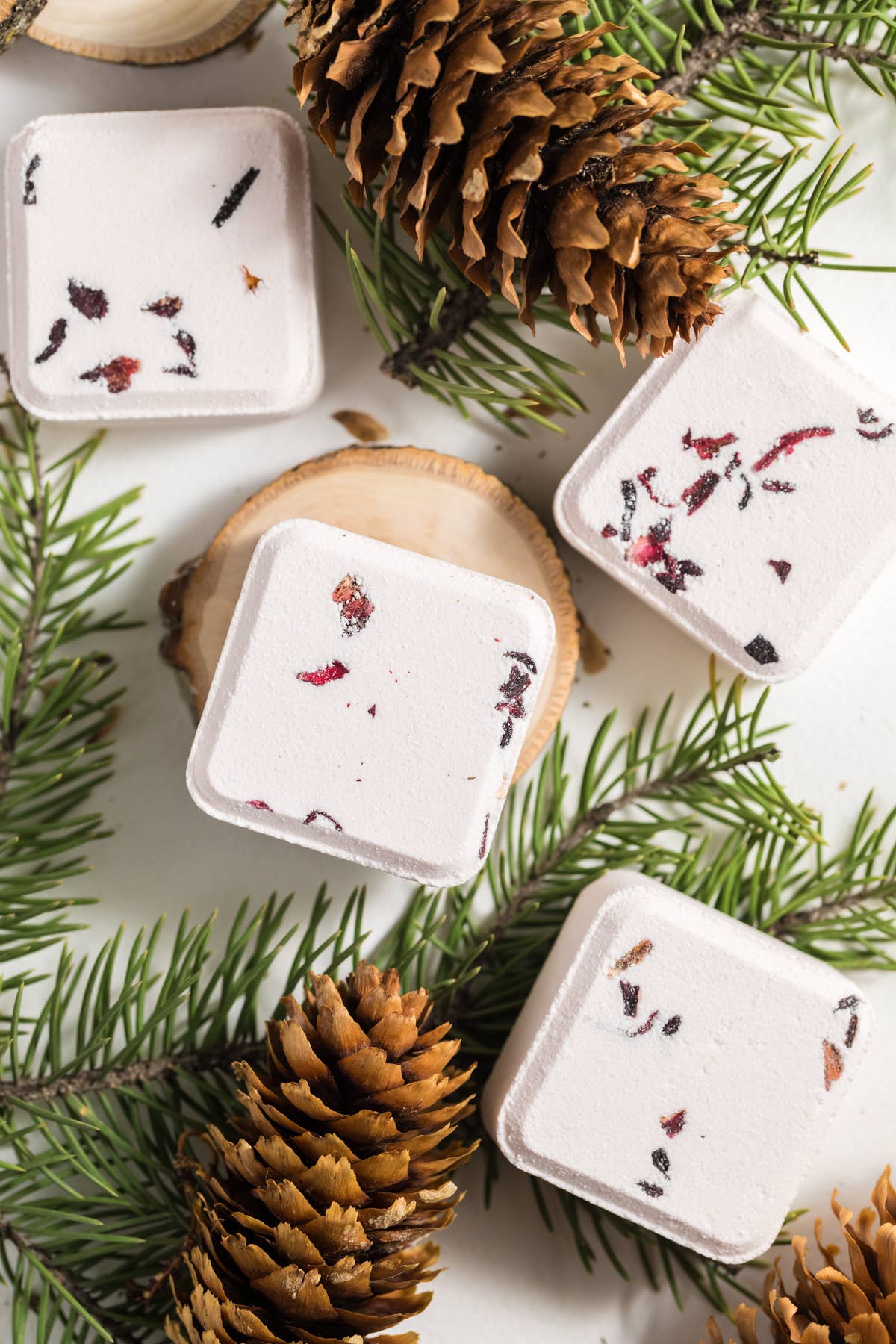 White shower steamers with dried herbs on a white surface with pine cones and branches.