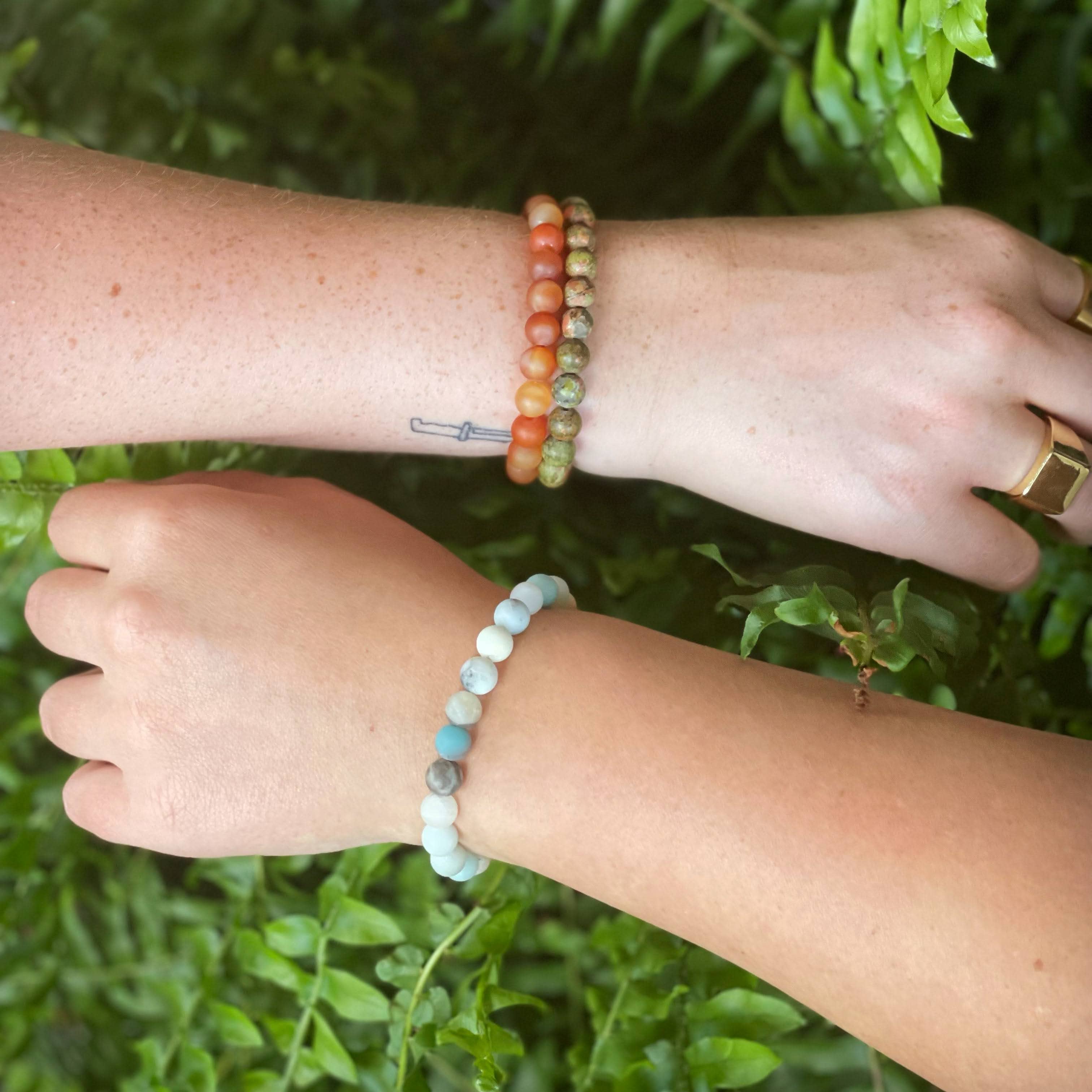 Two wrists wearing beaded bracelets against a green leafy background