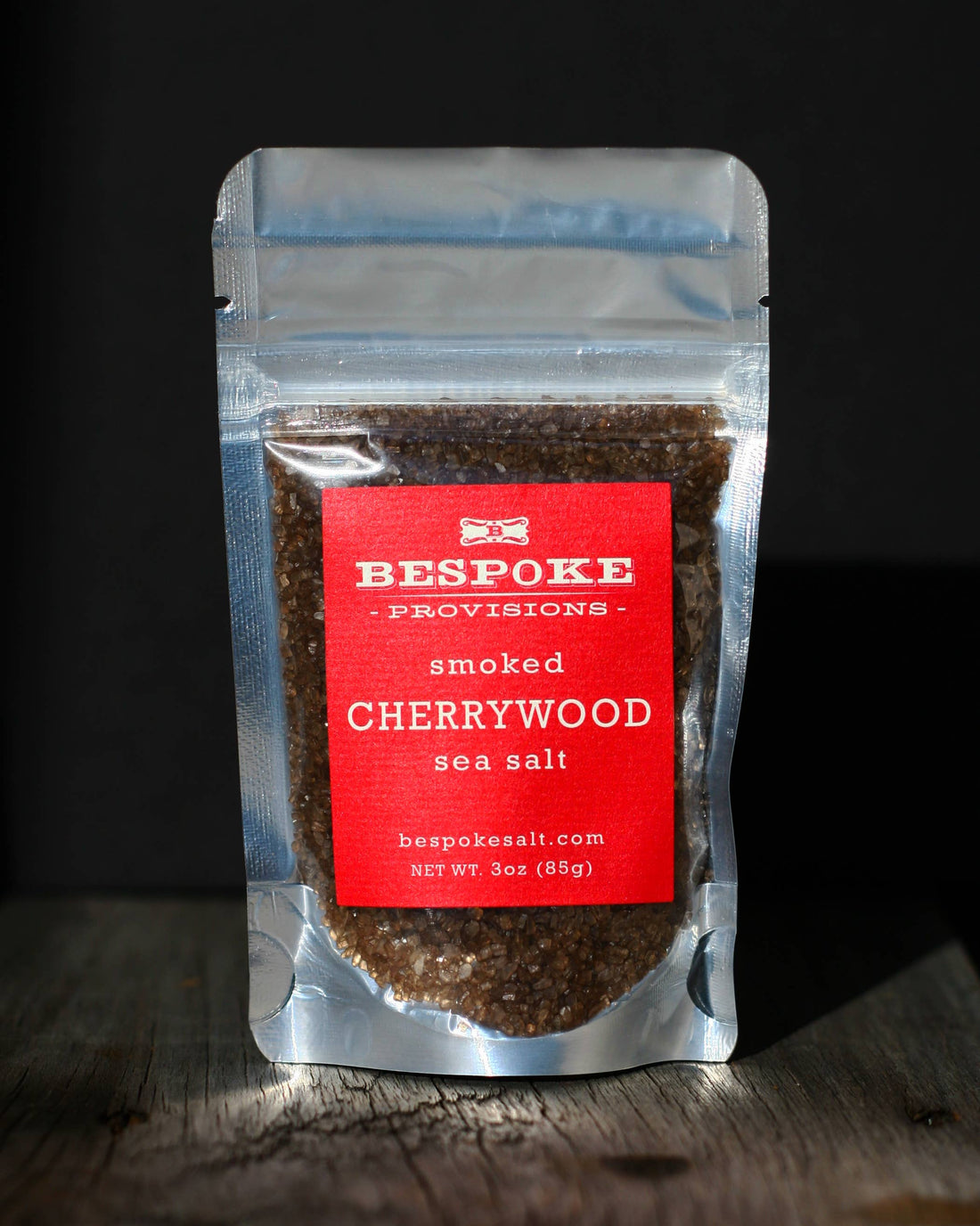A package of Bespoke Provisions smoked cherrywood sea salt placed on a wooden surface against a dark background.