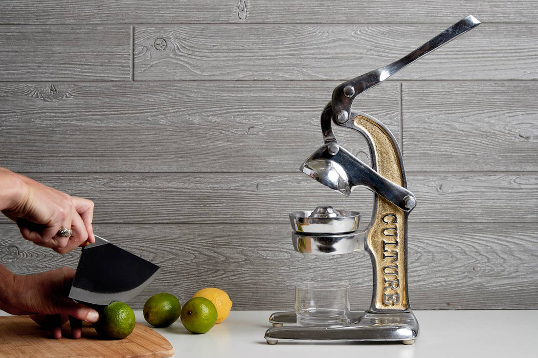 Vintage-style manual juicer on a wooden surface with limes and a knife against a wooden panel background.