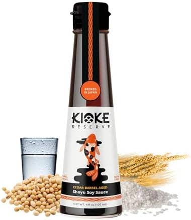 A bottle of Kioke Reserve Cedar Barrel Aged Shoyu Soy Sauce centered on a white background, with soybeans, a glass of water, and wheat stalks arranged around it.