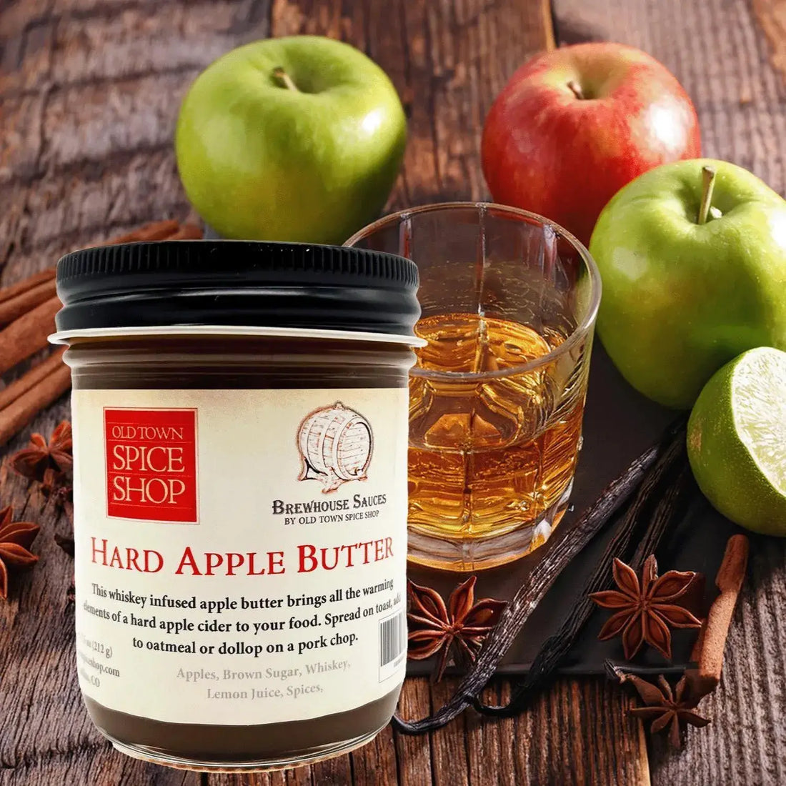 Jar of Old Town Spice Shop Hard Apple Butter with a glass of whiskey, apples, and spices on a wooden surface.