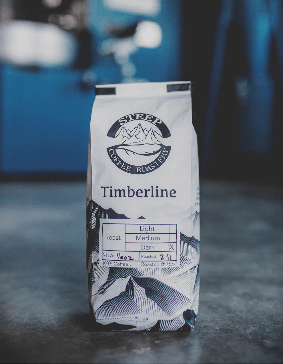 A bag of Silver Mountain Coffee Roasters Timberline light roast coffee stands on a dark surface with a blurred blue workshop background.