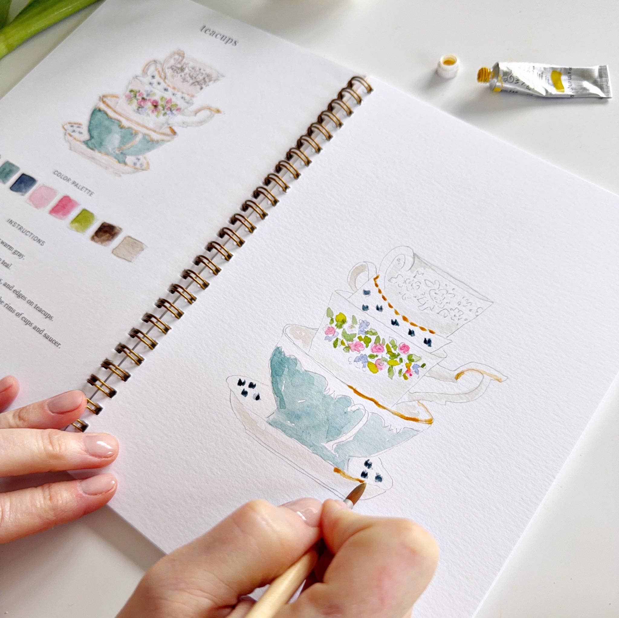Person drawing a teapot illustration in a sketchbook with paint tubes nearby.