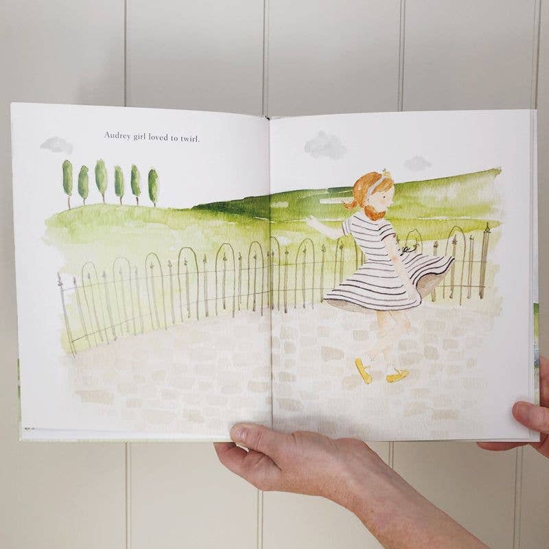 Illustrated book open to a page with a girl in a striped dress sitting on a fence.