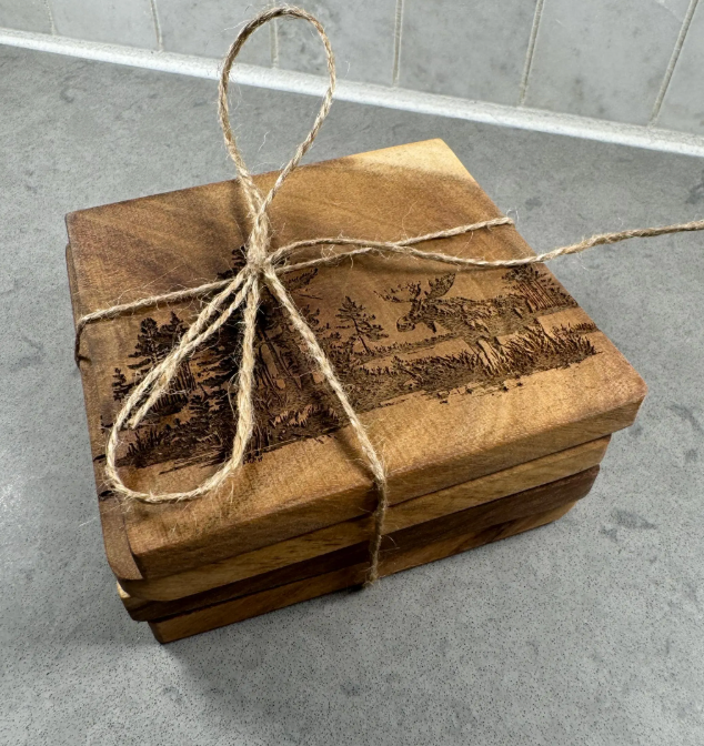 A stack of square wooden coasters with engraved landscape scenes, tied together with natural jute twine on a gray countertop.