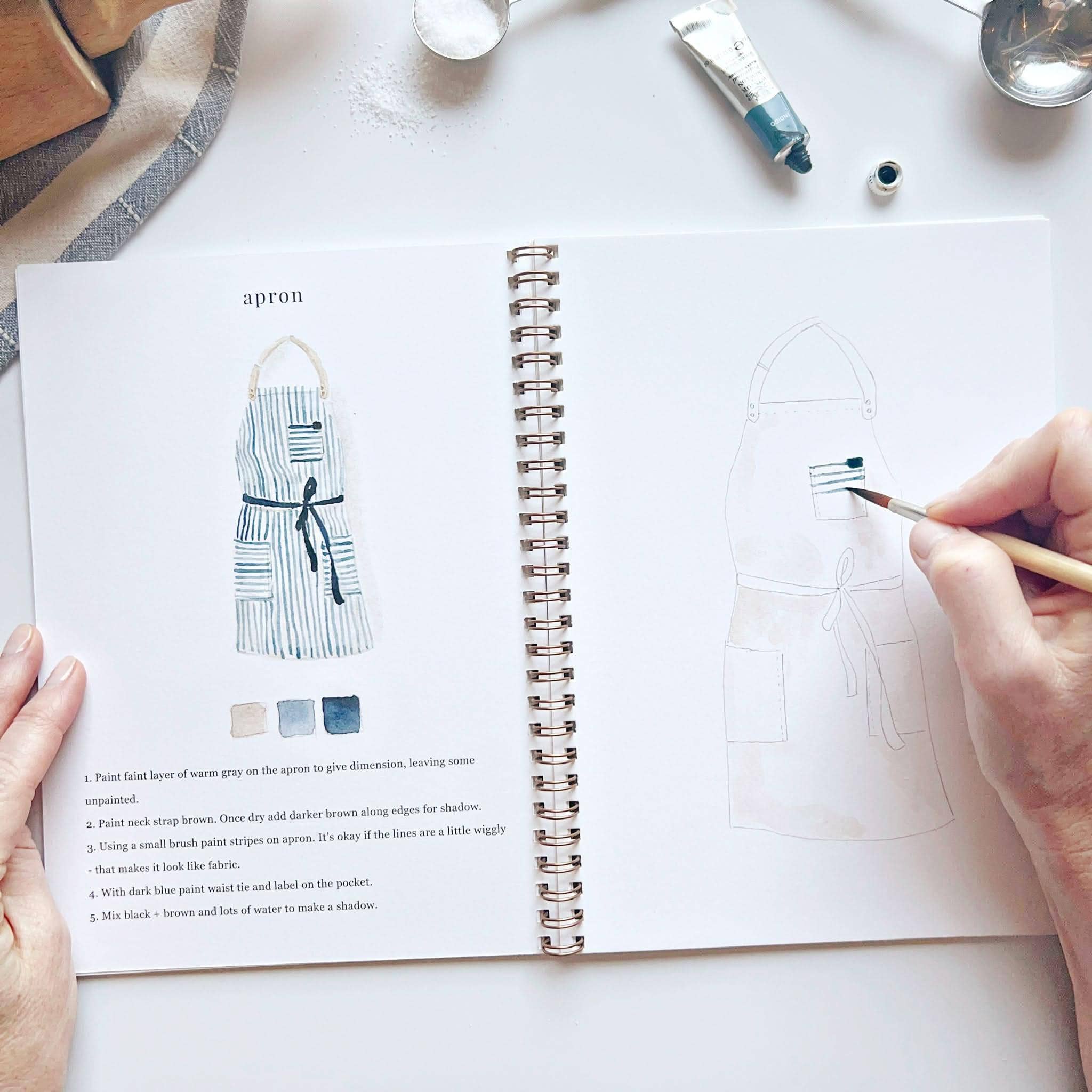 Person holding a paintbrush over an open book with apron design illustrations on a white surface.