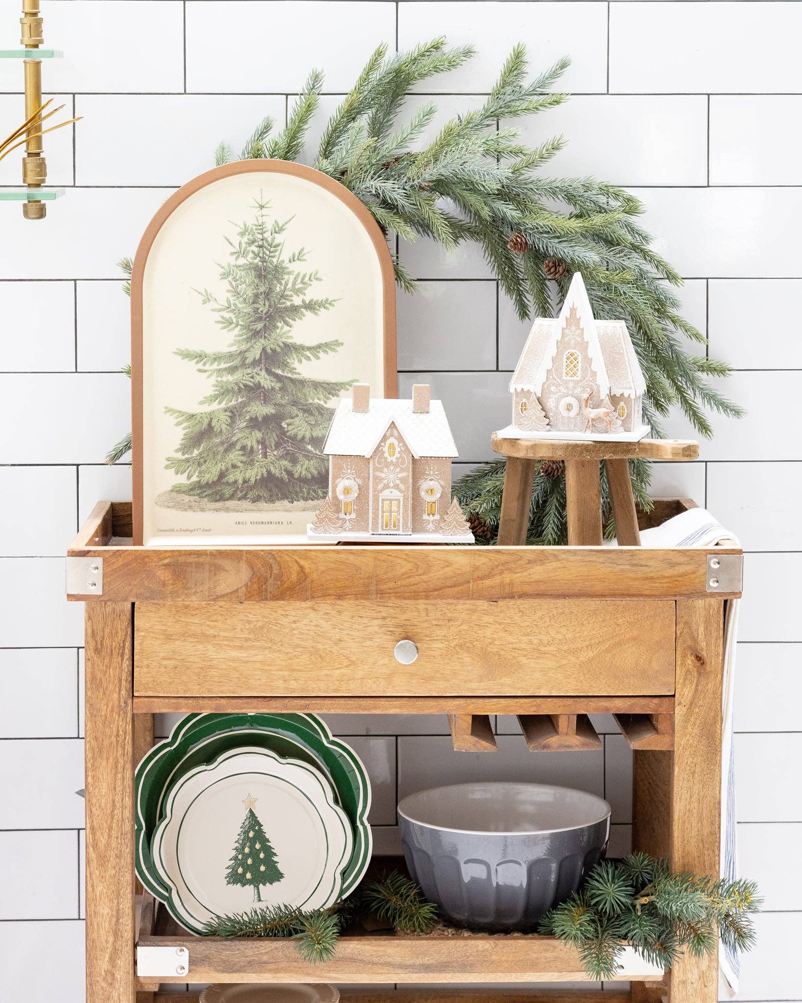 Wooden shelf with Christmas decor including a mirror, small houses, and greenery against a white tiled wall.
