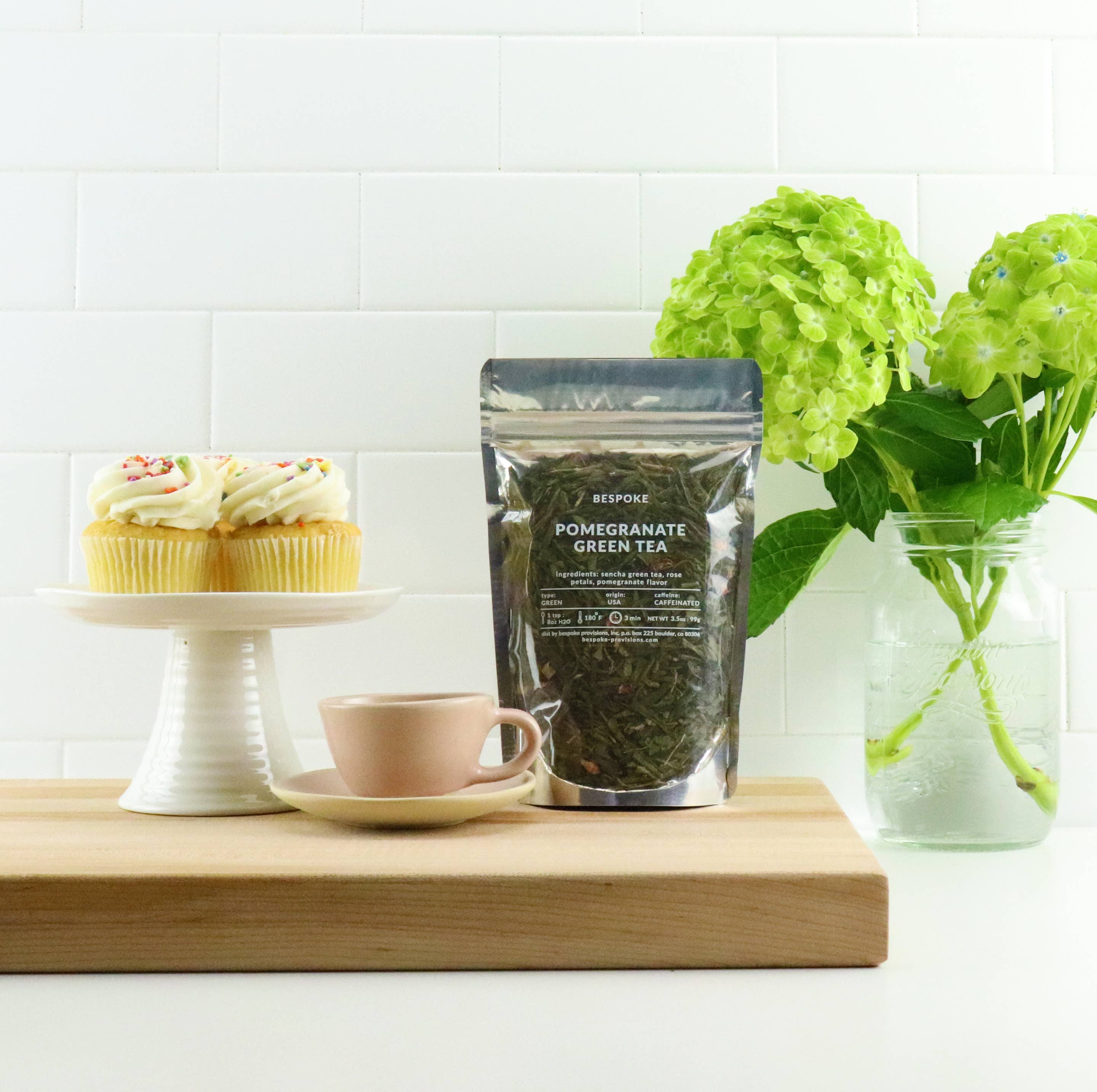 Pomegranate green tea package on a wooden board with a cup, saucer, and cupcake in the background.