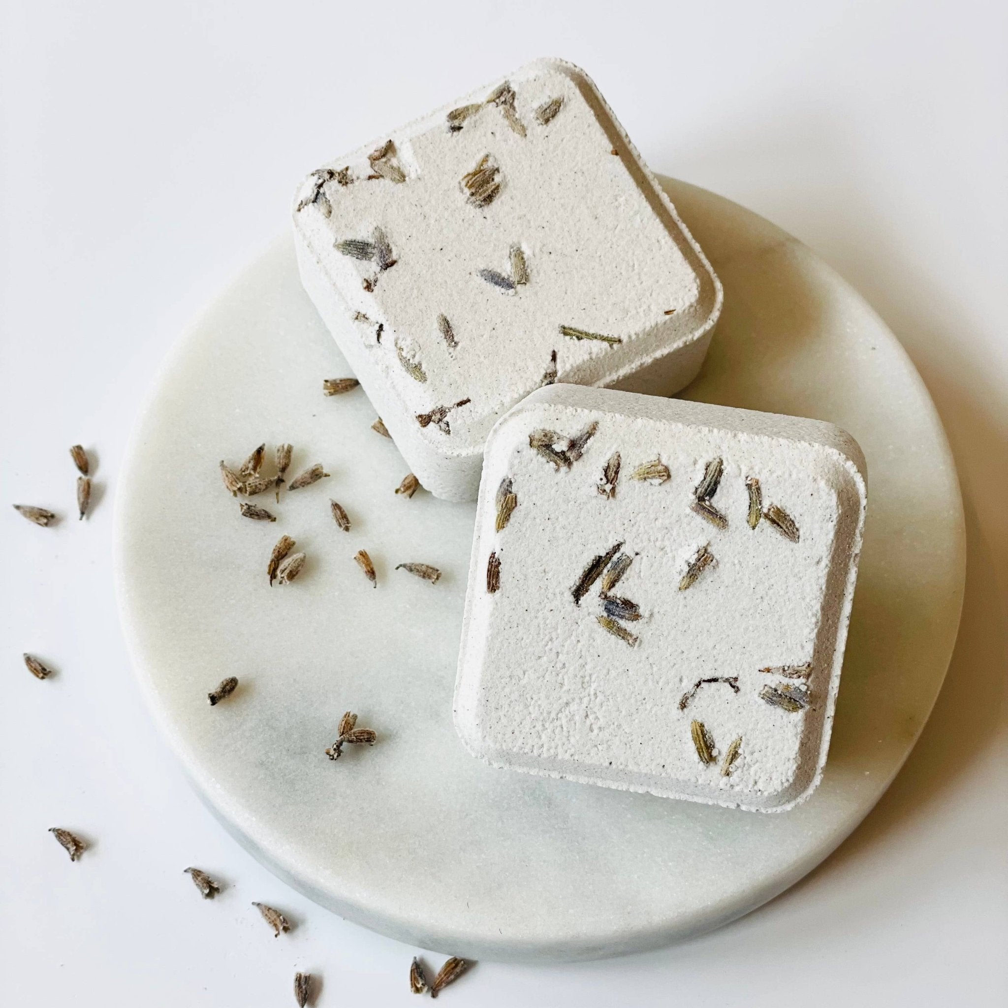 Two square shower steamers with dried herbs on a marble plate with scattered herbs on a light background