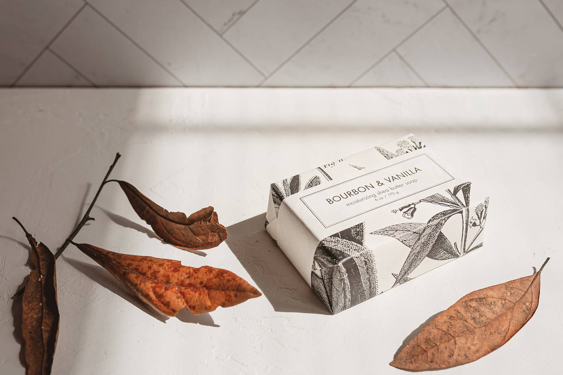 Bar of soap with packaging on a white surface with dried leaves and a branch.