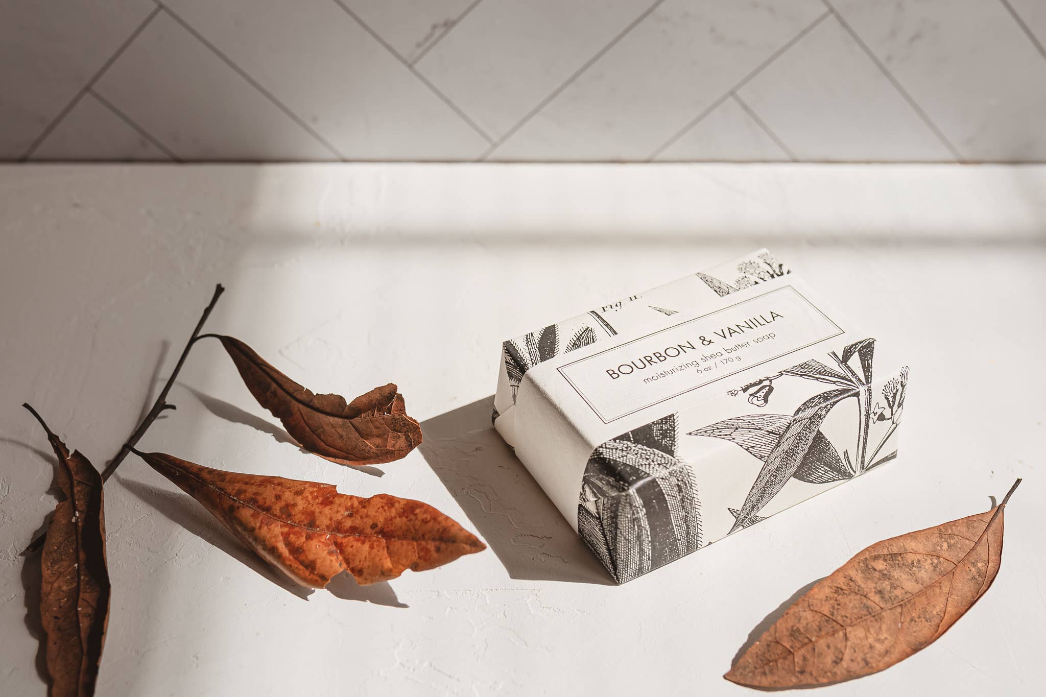 Bar of soap with packaging on a white surface with dried leaves and a branch.