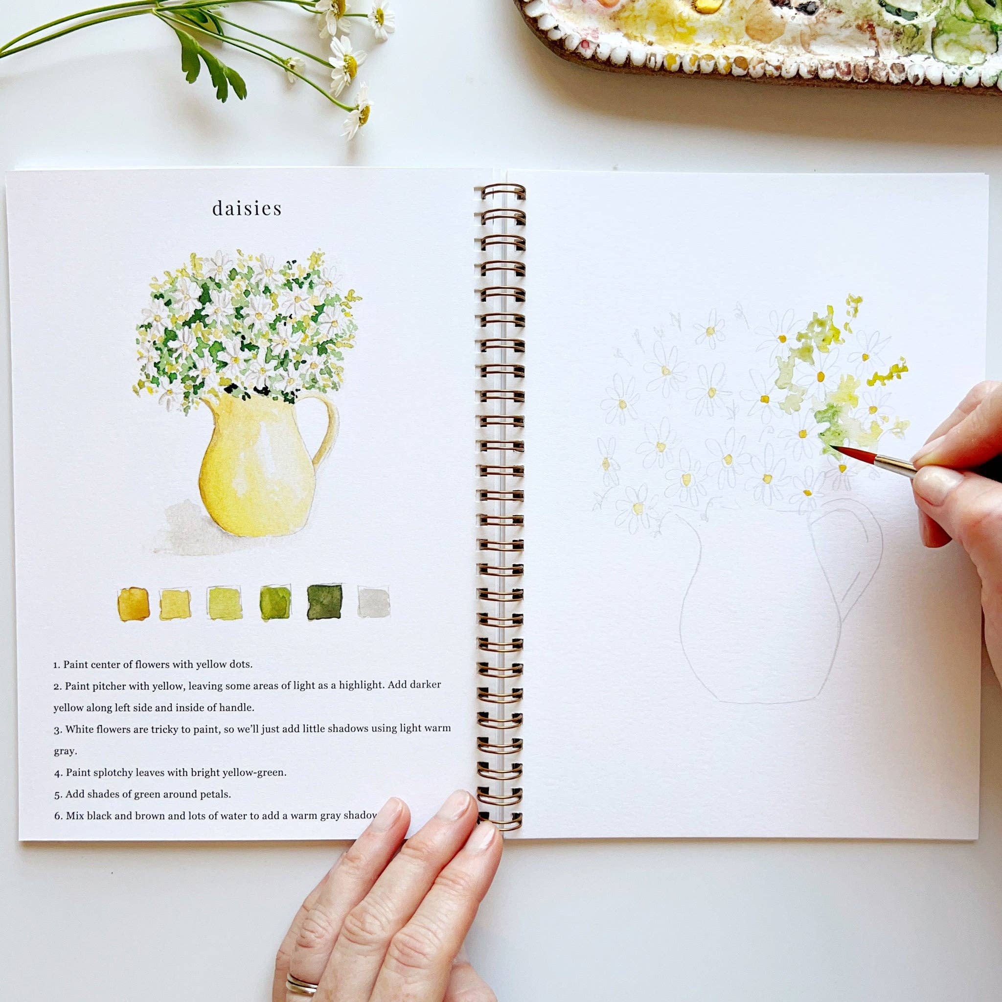 Open book with floral illustrations and a hand holding a paintbrush
