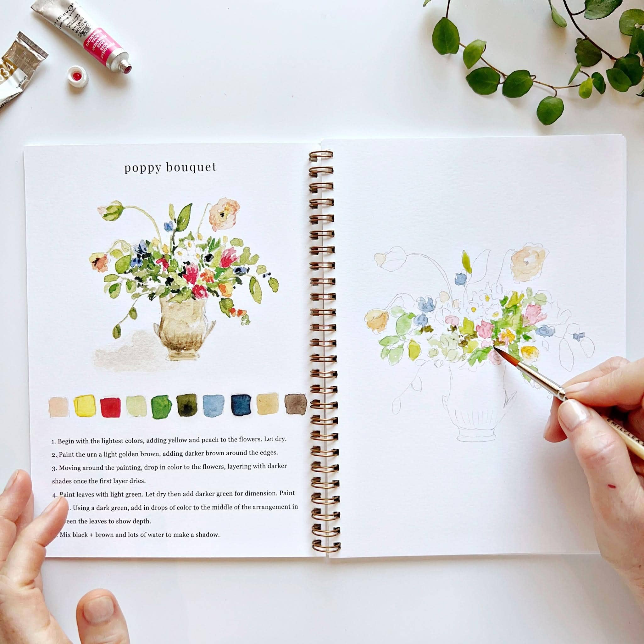 Open book with floral illustrations and color swatches, held by a person with a paintbrush.