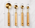 Set of four gold measuring spoons with wooden handles, each labeled with dimensions and weight.