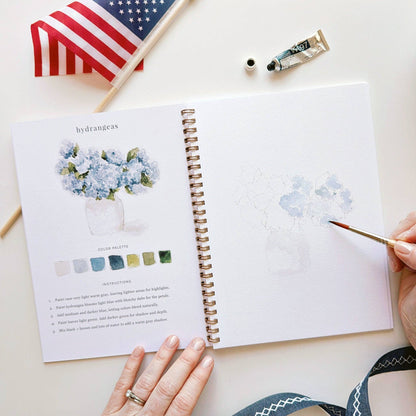 Open notebook with watercolor painting and color palette on a white surface with a small American flag.