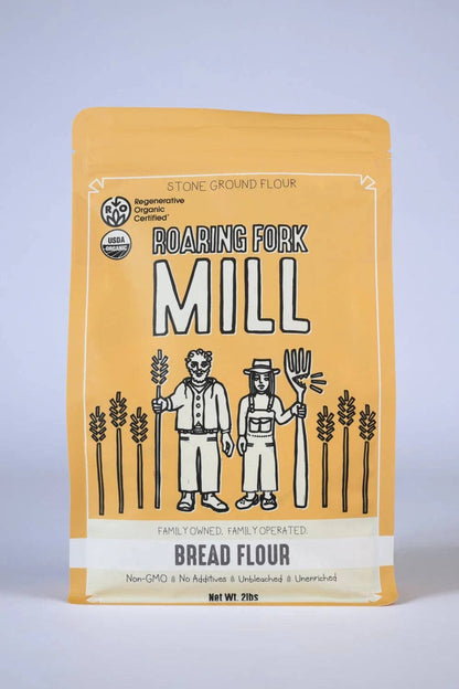 A stand-up pouch of Roaring Fork Mill Bread Flour on a light gray studio background. The package is predominantly warm yellow-orange with a cream lower section and black-and-white illustrated figures and wheat stalks.