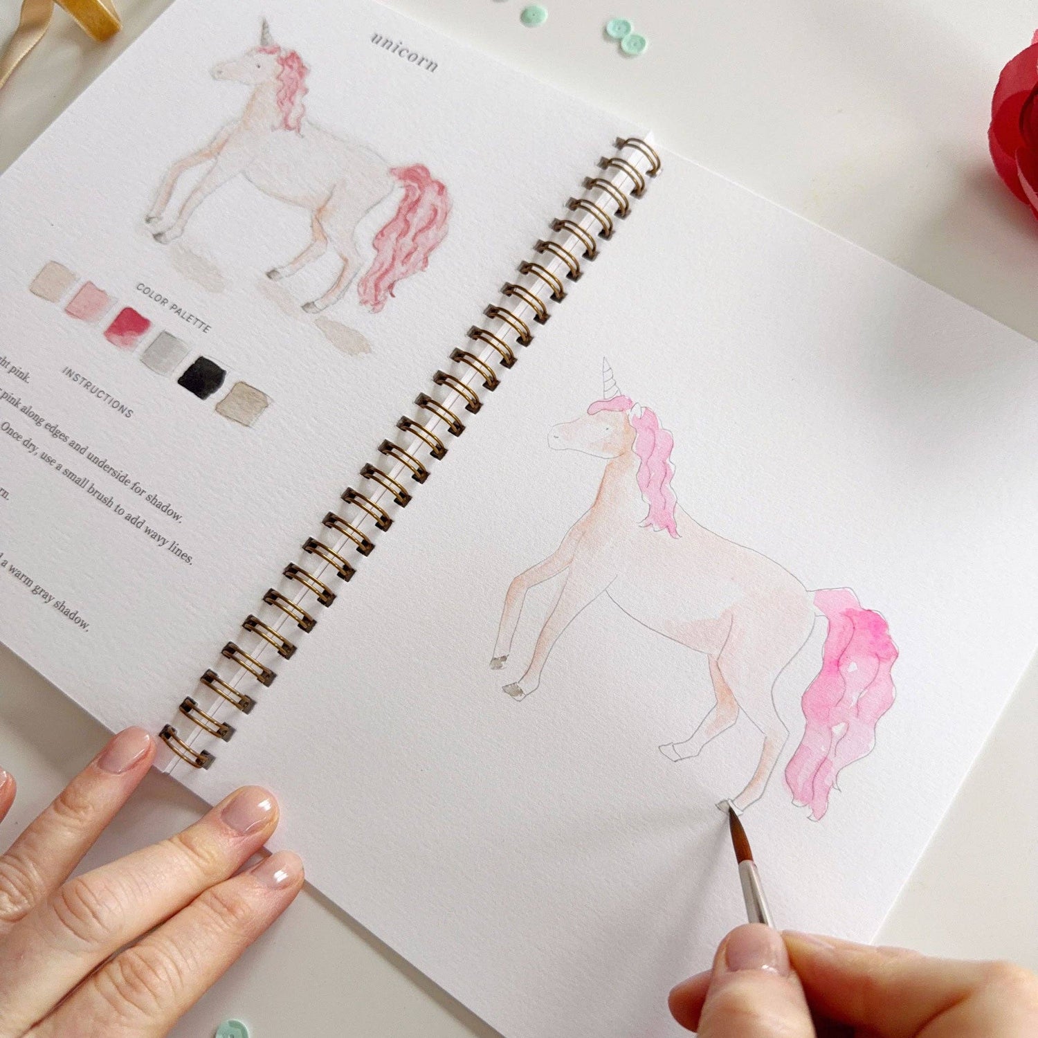 Person coloring a unicorn illustration in a sketchbook with color swatches.