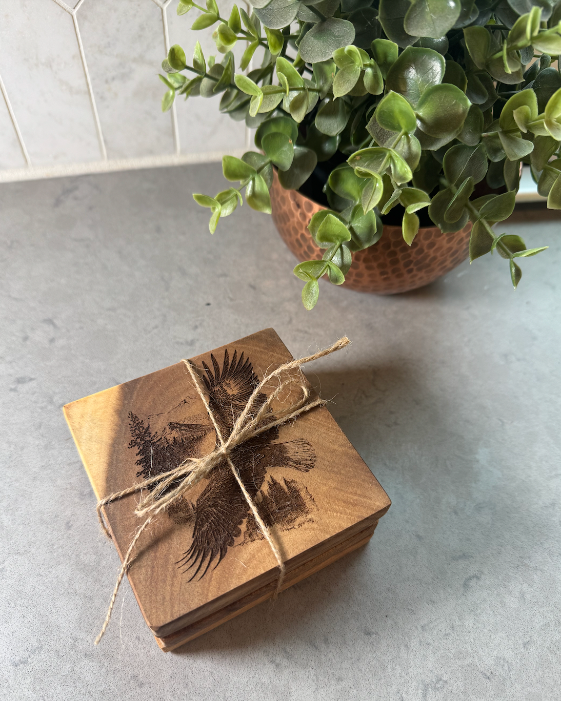 A stack of square wooden coasters tied with twine, featuring a dark engraved eagle design on the top piece, set on a gray countertop with a potted green plant in the background.