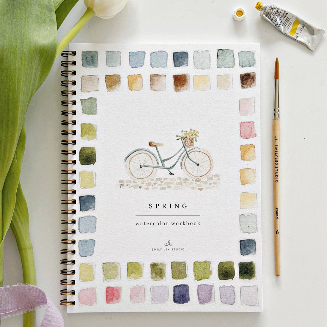 Spring watercolor workbook with a bicycle illustration and color swatches, surrounded by art supplies on a white surface.