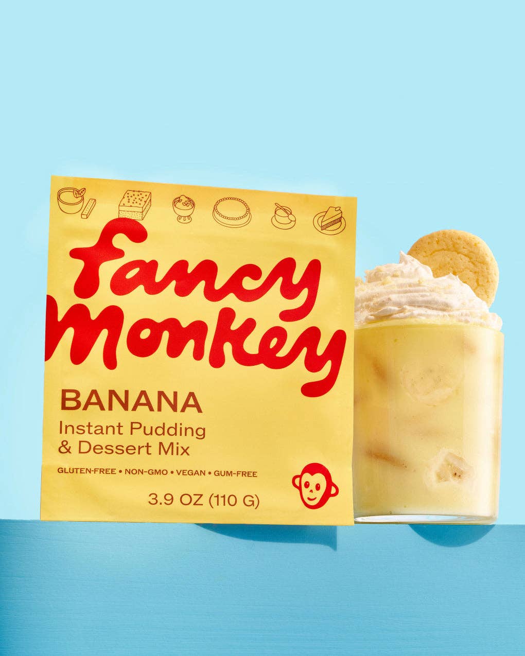 A yellow box of Fancy Monkey Banana Instant Pudding &amp; Dessert Mix stands next to a glass of creamy banana pudding topped with whipped cream and a cookie, set against a light blue background.