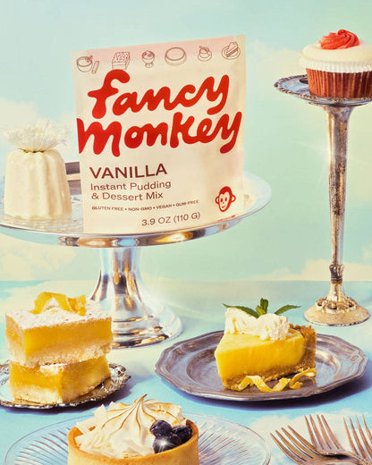 A package of Fancy Monkey Vanilla Instant Pudding and Dessert Mix stands on a silver cake stand, surrounded by styled dessert slices and a cupcake on plates against a pale blue background.