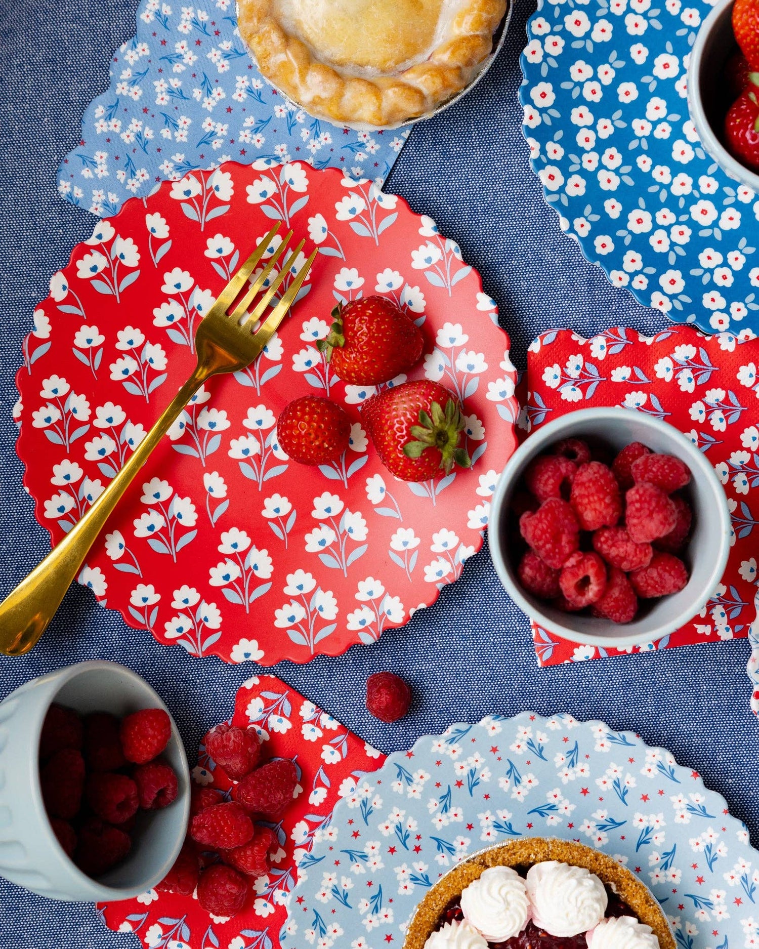 Table setting with red and blue floral plates, raspberries, and a pie on a blue tablecloth.