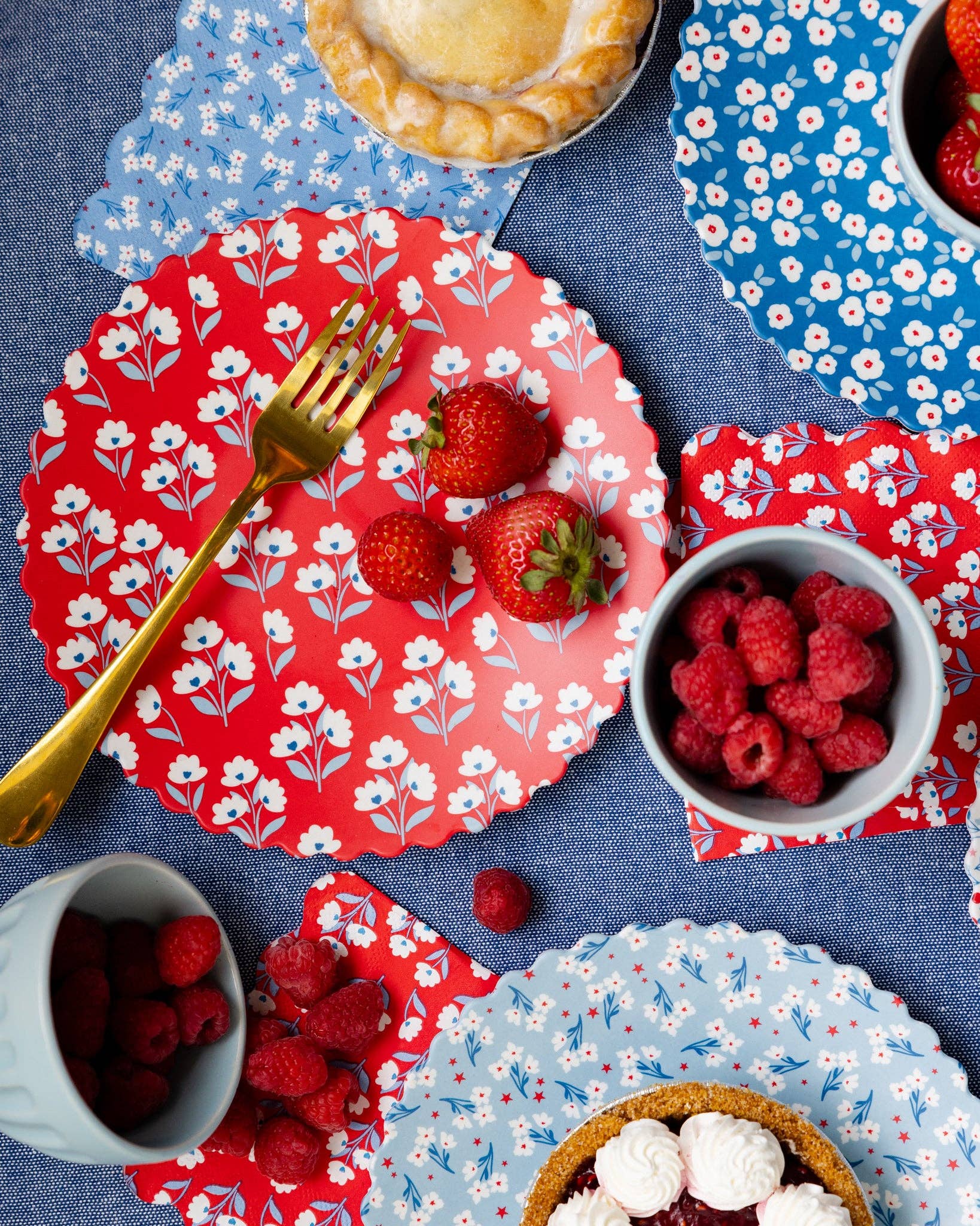 Table setting with red and blue floral plates, raspberries, and a pie on a blue tablecloth.