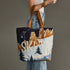 A person wearing light-wash blue jeans and a white top carries a large canvas tote bag with brown leather handles. The bag features a stylized winter mountain landscape print with the words &