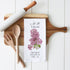 A white kitchen tea towel with a purple lilac floral print and elegant black typography, styled on a wooden cutting board with a rolling pin, wooden utensils, and a white paneled background.