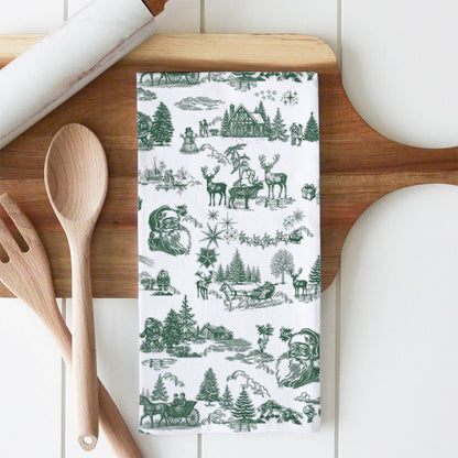 A white kitchen towel with a green Christmas-themed toile de Jouy pattern featuring Santa Claus, reindeer, sleighs, and trees, styled on a wooden cutting board with wooden spoons and a rolling pin.