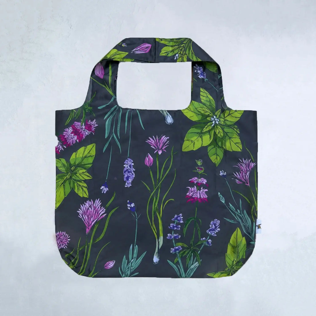 A reusable tote bag with a dark charcoal background and a vibrant botanical print featuring green leaves, purple wildflowers, and blue-green stems, shown against a light gray textured background.
