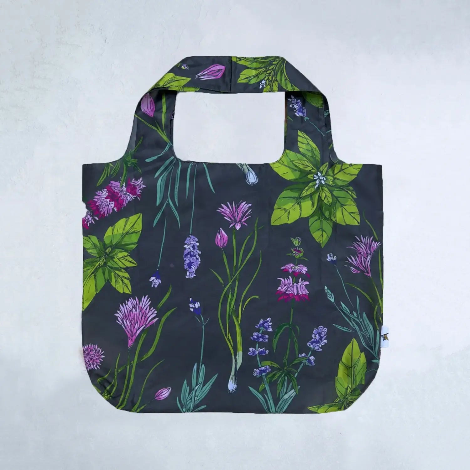 A reusable tote bag with a dark charcoal background and a vibrant botanical print featuring green leaves, purple wildflowers, and blue-green stems, shown against a light gray textured background.