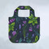 A reusable tote bag with a dark charcoal background and a vibrant botanical print featuring green leaves, purple wildflowers, and blue-green stems, shown against a light gray textured background.