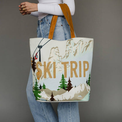 A person wearing a white long-sleeve top and light blue jeans holds a large canvas tote bag printed with a ski mountain scene and the words “SKI TRIP” in tan. The bag has tan shoulder straps and a white base with tan lettering, brown trees, and gray mountain peaks.