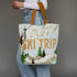 A person wearing a white long-sleeve top and light blue jeans holds a large canvas tote bag printed with a ski mountain scene and the words “SKI TRIP” in tan. The bag has tan shoulder straps and a white base with tan lettering, brown trees, and gray mountain peaks.