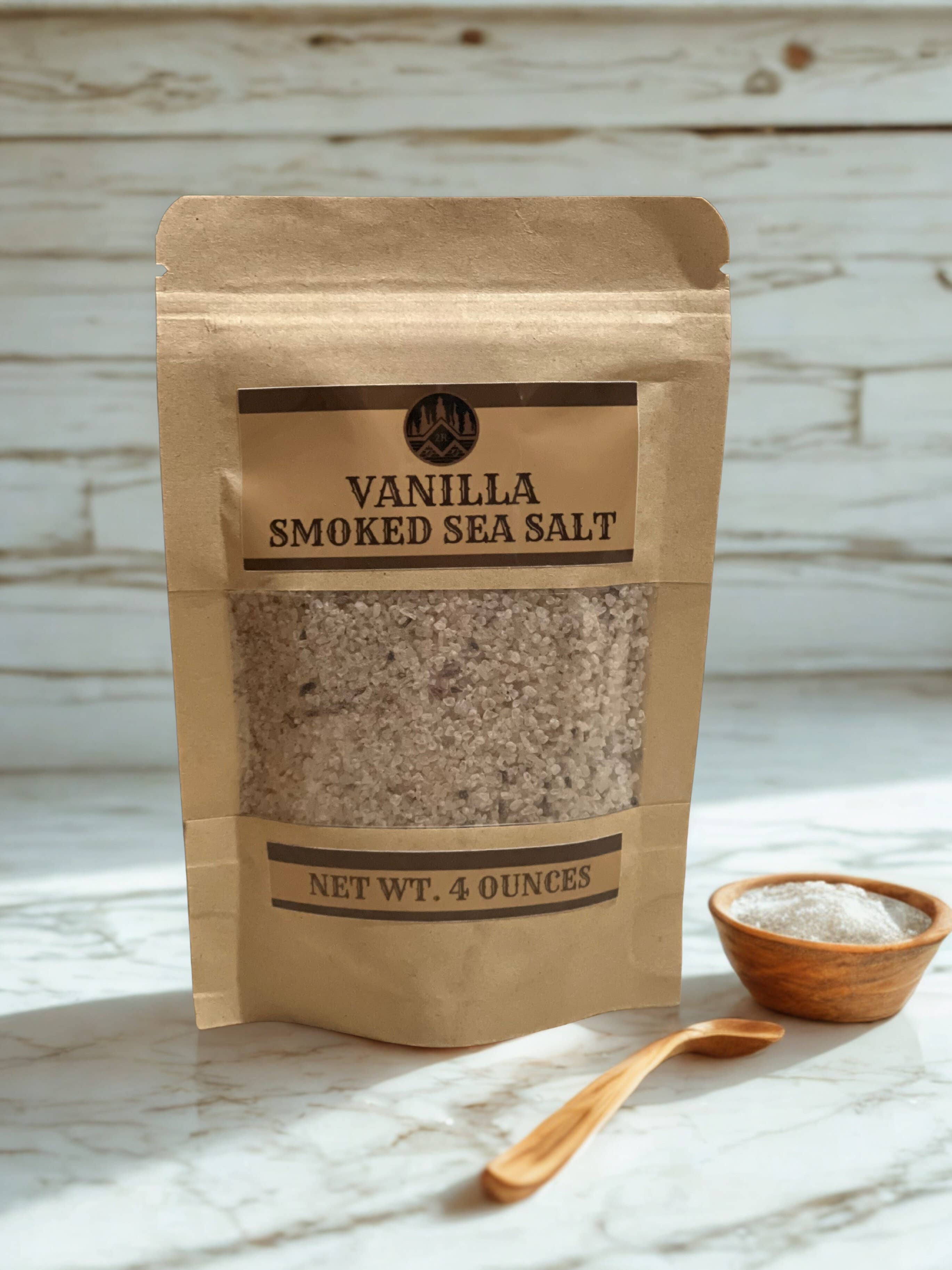 Kraft paper packaging of vanilla smoked sea salt on a marble surface with a wooden spoon and small bowl.