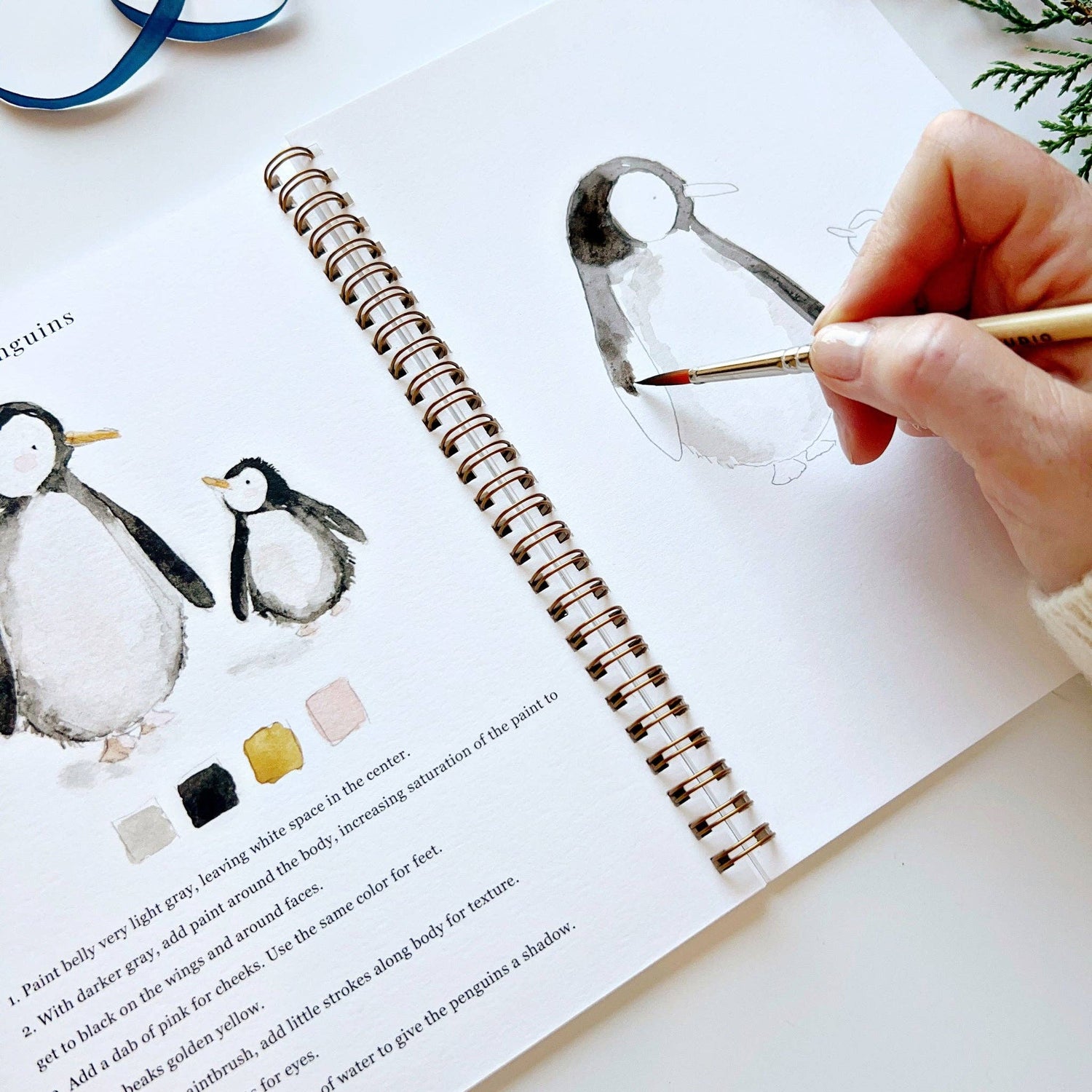 Person painting a penguin illustration with watercolors on a book open to a page with penguin illustrations.