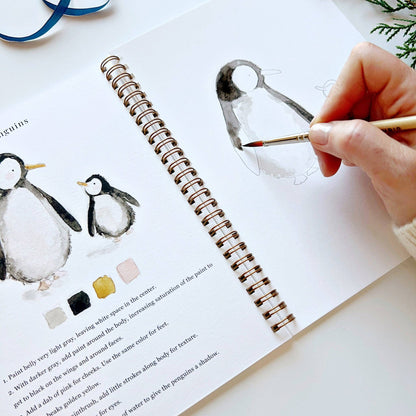 Person painting a penguin illustration with watercolors on a book open to a page with penguin illustrations.