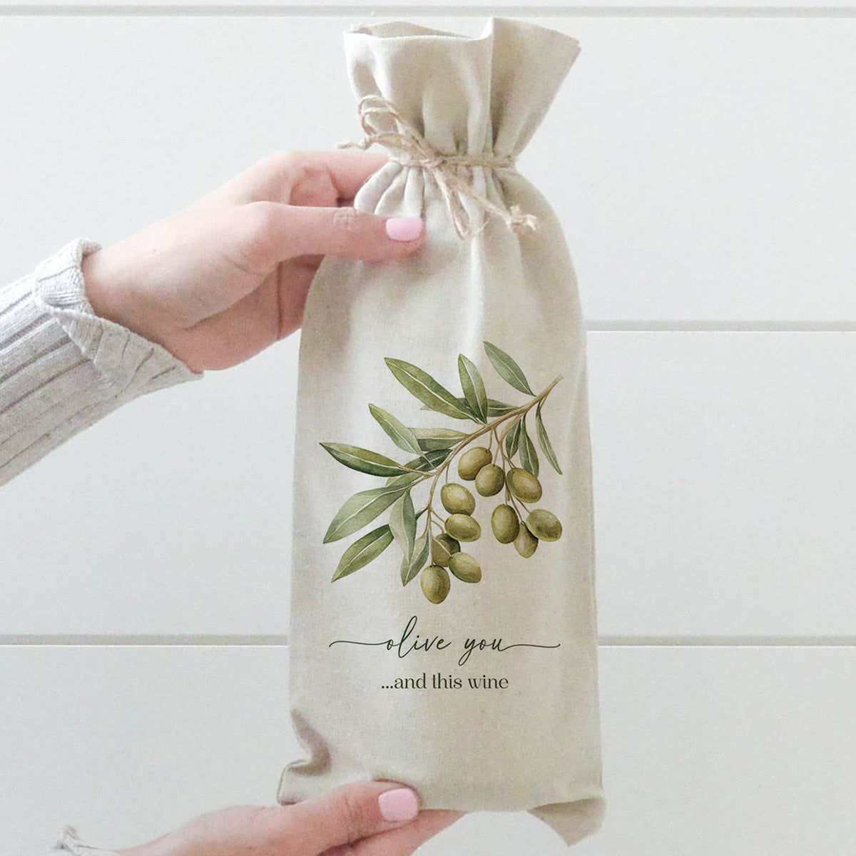 A person holds a beige fabric wine bag with a watercolor olive branch illustration and the text &