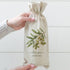 A person holds a beige fabric wine bag with a watercolor olive branch illustration and the text &