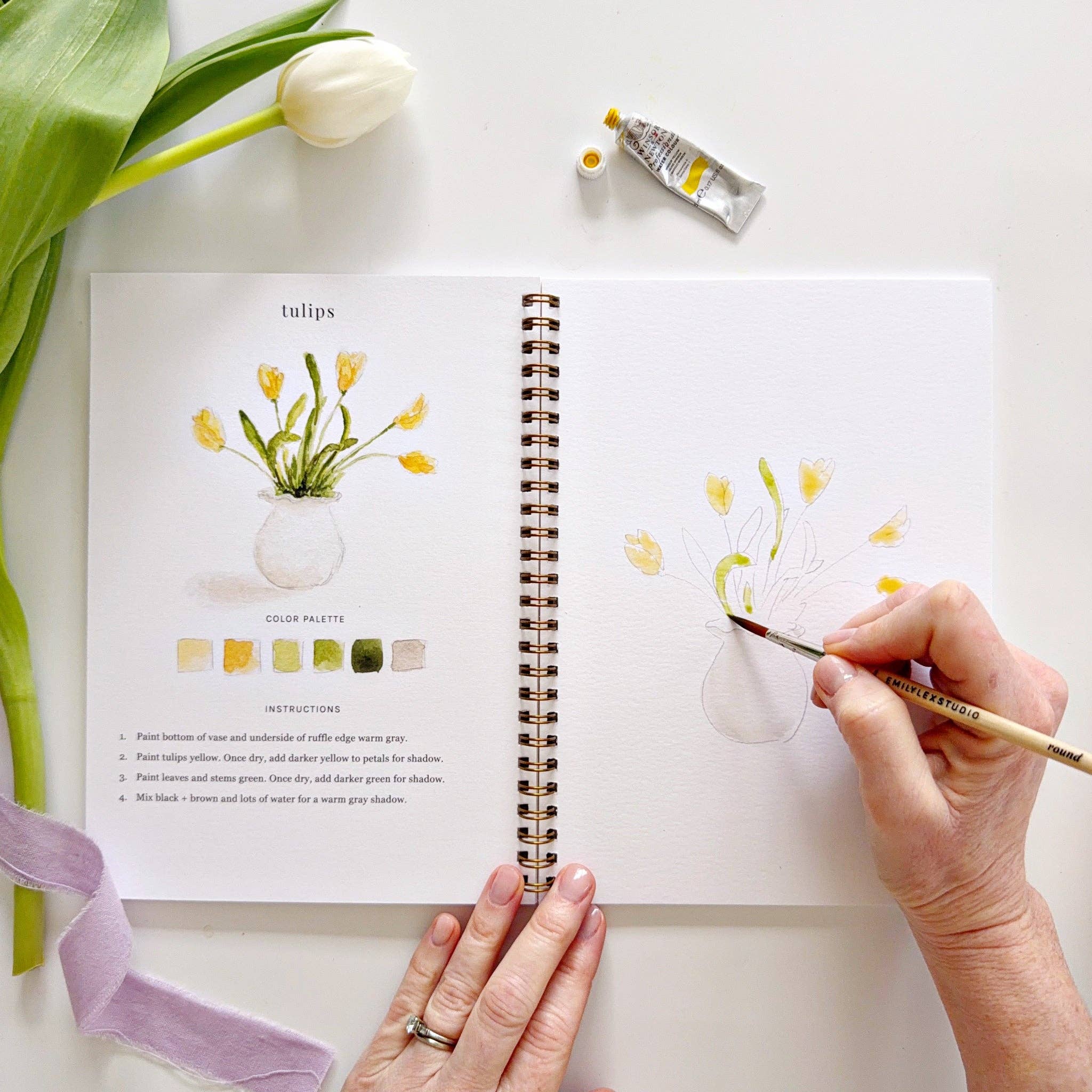 Person painting tulips on a sketchbook with a bottle of water and a tulip flower nearby.