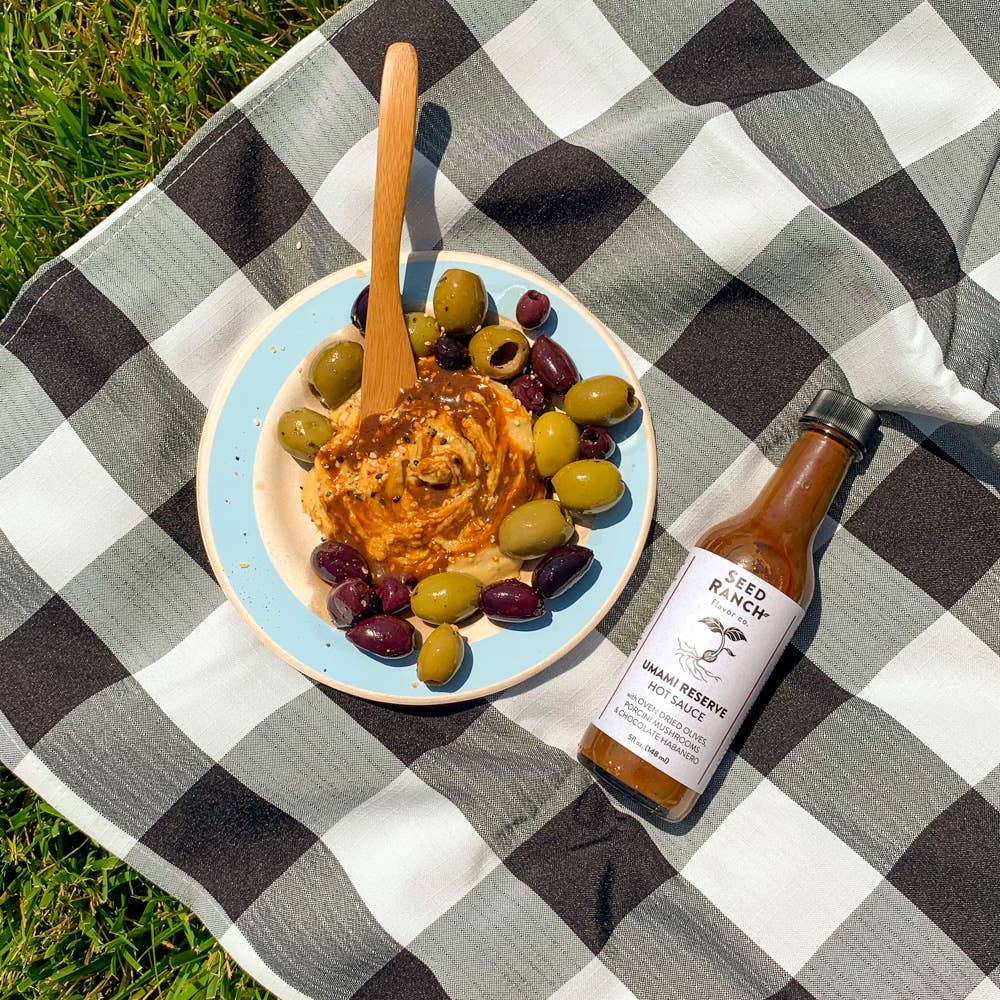 Dip with olives and a bottle of sauce on a checkered blanket outdoors