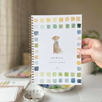 Hand holding a watercolor workbook with a dog illustration on a blurred indoor background